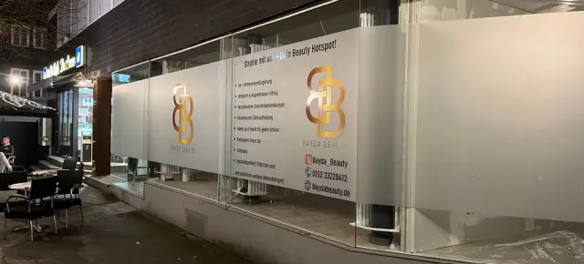 Glasfront von Bayda Beauty mit Sichtschutzfolie, goldenem Logo und Leistungsliste, abends beleuchtet
